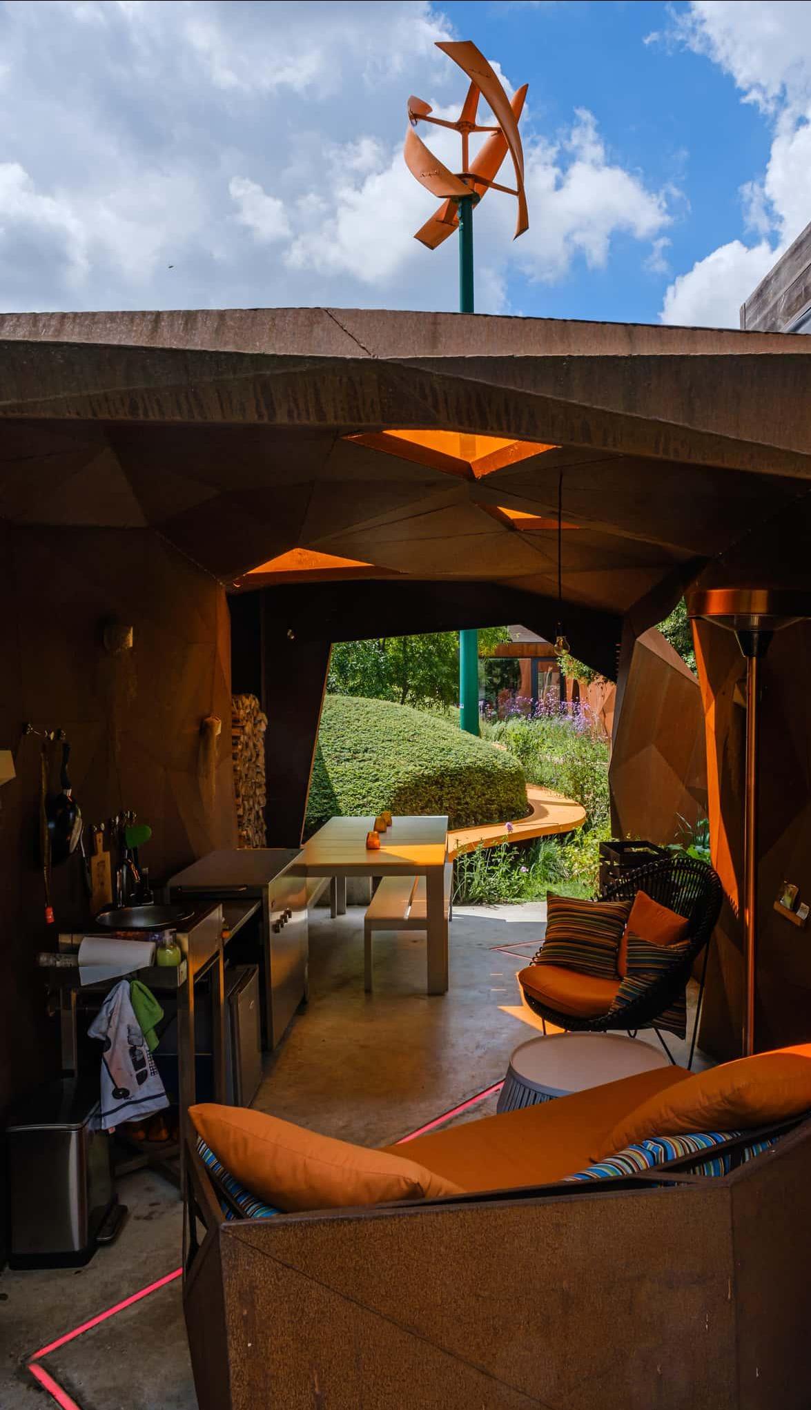 De beweging in metaal wordt een sculptuur die zich een weg baant door de tuin en achter de windmolen een shelter vormt, waar men zich kan terugtrekken.nGekoppeld aan de shelter vinden we een leuk vormgegeven moestuin terug met leifruit.nHier kan men de vrucht van de dag plukken en onmiddellijk in de kleine keuken klaarmaken tot een lekker gerecht.n